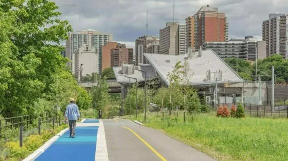 Voie cyclable et sentier piétonnier séparés. Des personnes marchent sur le sentier piétonnier; la station Pimisi de l’O-Train est en arrière-plan.