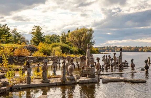 Approximately thirty balanced rock sculptures on the empty shoreline of a wide river, in fall.