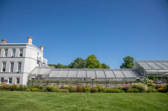 Ceremonial greenhouse attached to the main Rideau Hall building, surrounded by manicured lawns, colourful flower beds, and trees under a clear blue sky.
