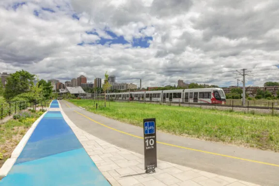 Voie cyclable et sentier piétonnier séparés, avec signalisation de la vitesse (maximum 10 km/h), et la station de l’O-Train Pimisi en arrière-plan.