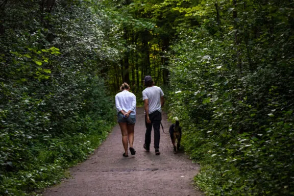 Deux adultes marchent avec un chien sur un tronçon de sentier plat, en poussière de pierre, au cœur d’une forêt dense.