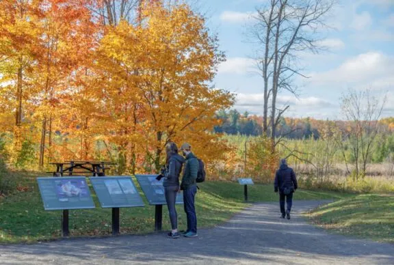 Un large sentier en poussière de pierre compactée avec une légère pente descendante. Des gens marchent et consultent des panneaux d’interprétation.