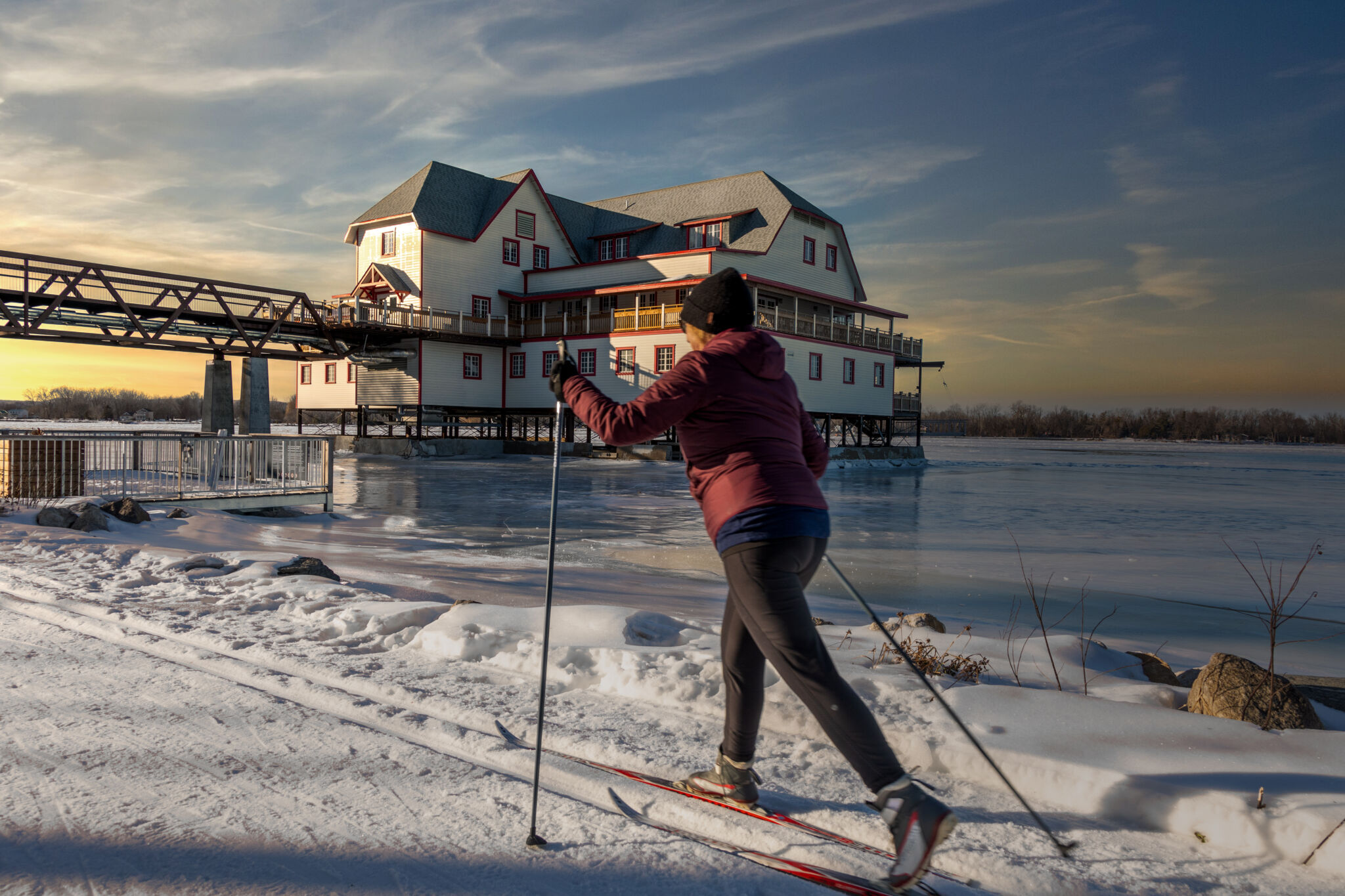 A person on cross-country skis skies along the frozen Ottawa River with the NCC River House in the background.