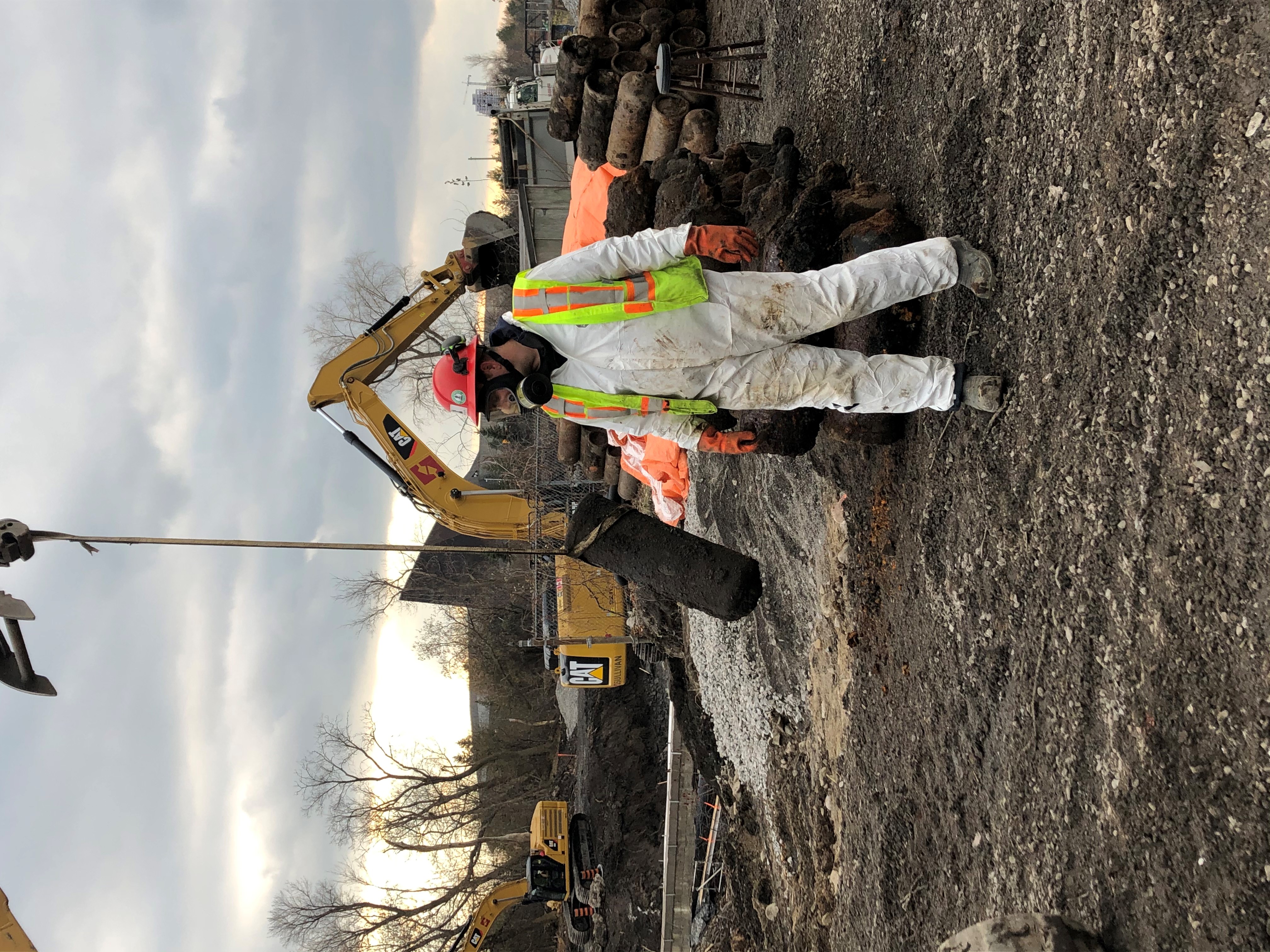 A worker geared up in a carbon-lined suit and full-face respirator stands in front of a pile of acetylene gas cylinders. In the background, there are mechanical excavators.