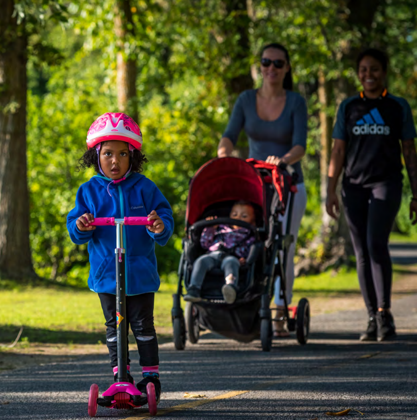 Deux personnes qui marchent côte à côte sur le Sentier de la capitale avec une poussette, derrière un enfant en trottinette.