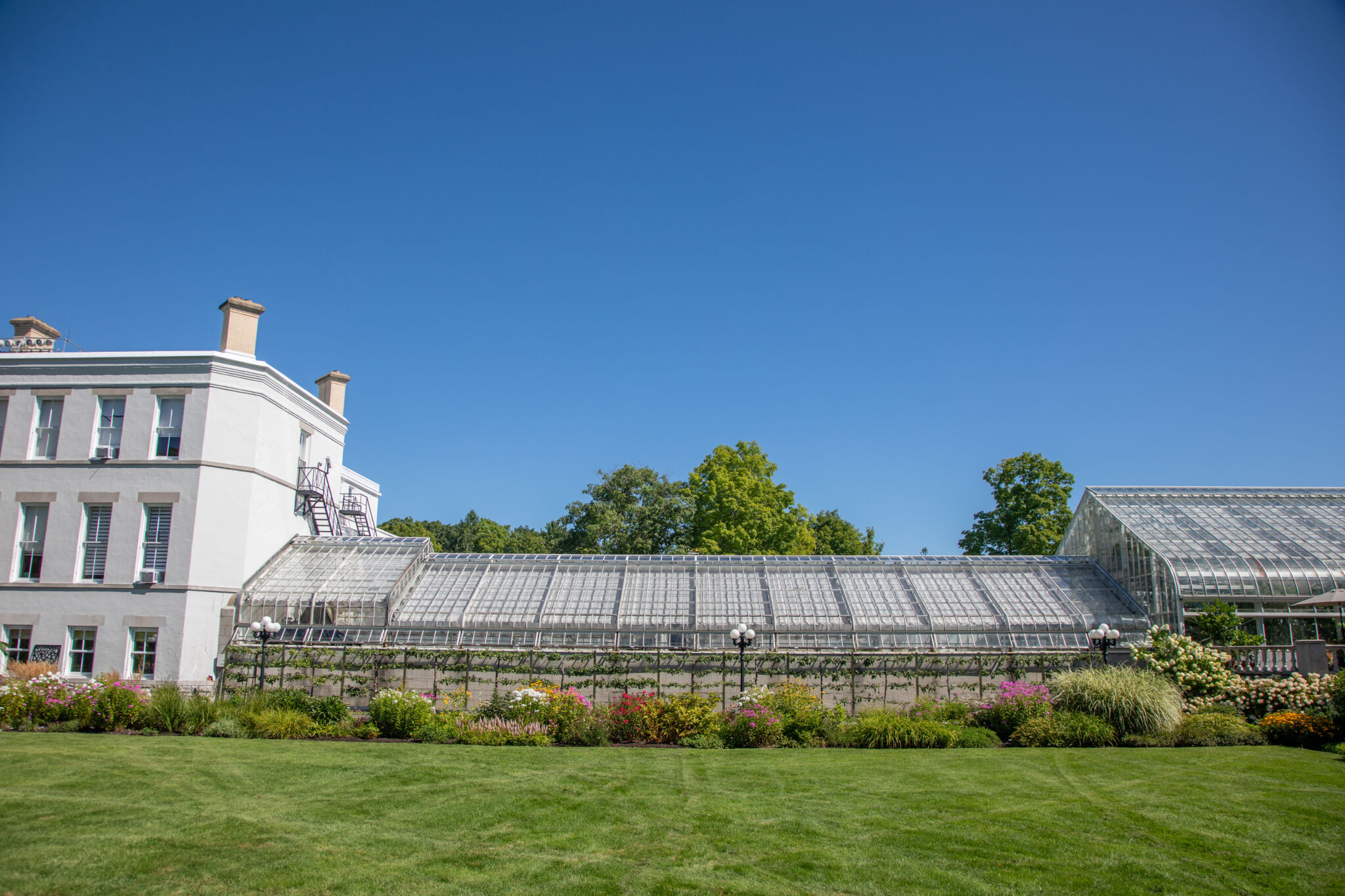 La serre d’exposition reliée au bâtiment principal de Rideau Hall entourée de pelouses entretenues, de parterres de fleurs colorés et d’arbres, sous un ciel bleu.