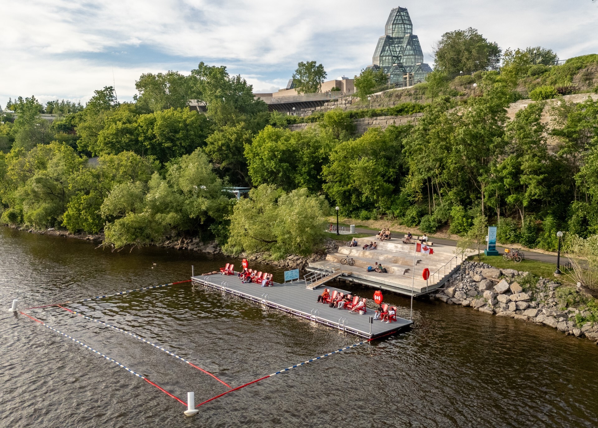 Un quai en bois, sur lequel sont disposées des chaises Muskoka rouges, et une vue sur des berges bordées d’arbres. À l’arrière-plan, la tour en verre du Musée des beaux-arts du Canada se dresse au-dessus de la végétation environnante.