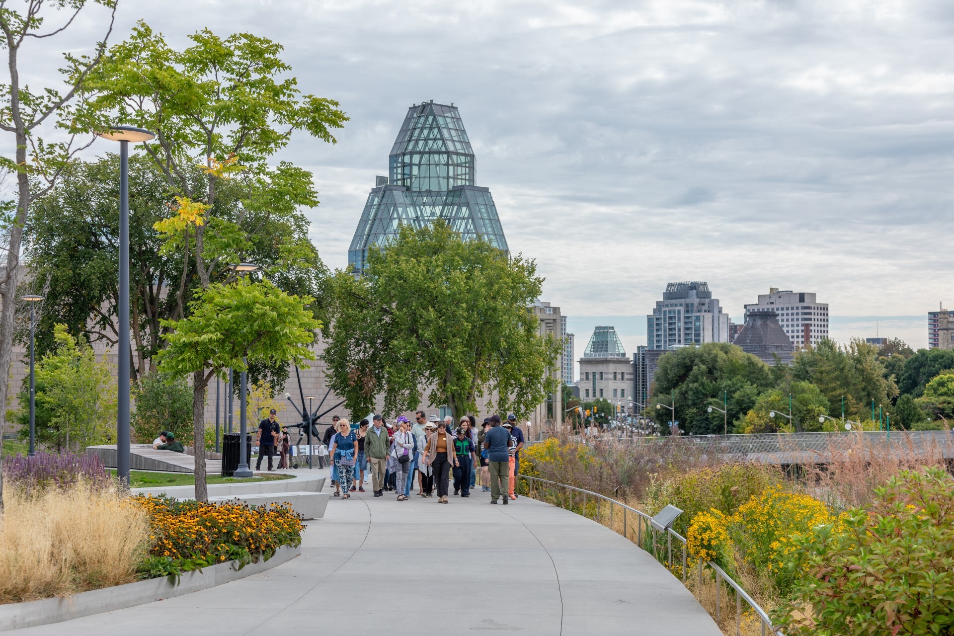Un groupe de personnes marche dans un cadre de verdure luxuriante. La tour en verre du Musée des beaux-arts du Canada se profile en arrière-plan.