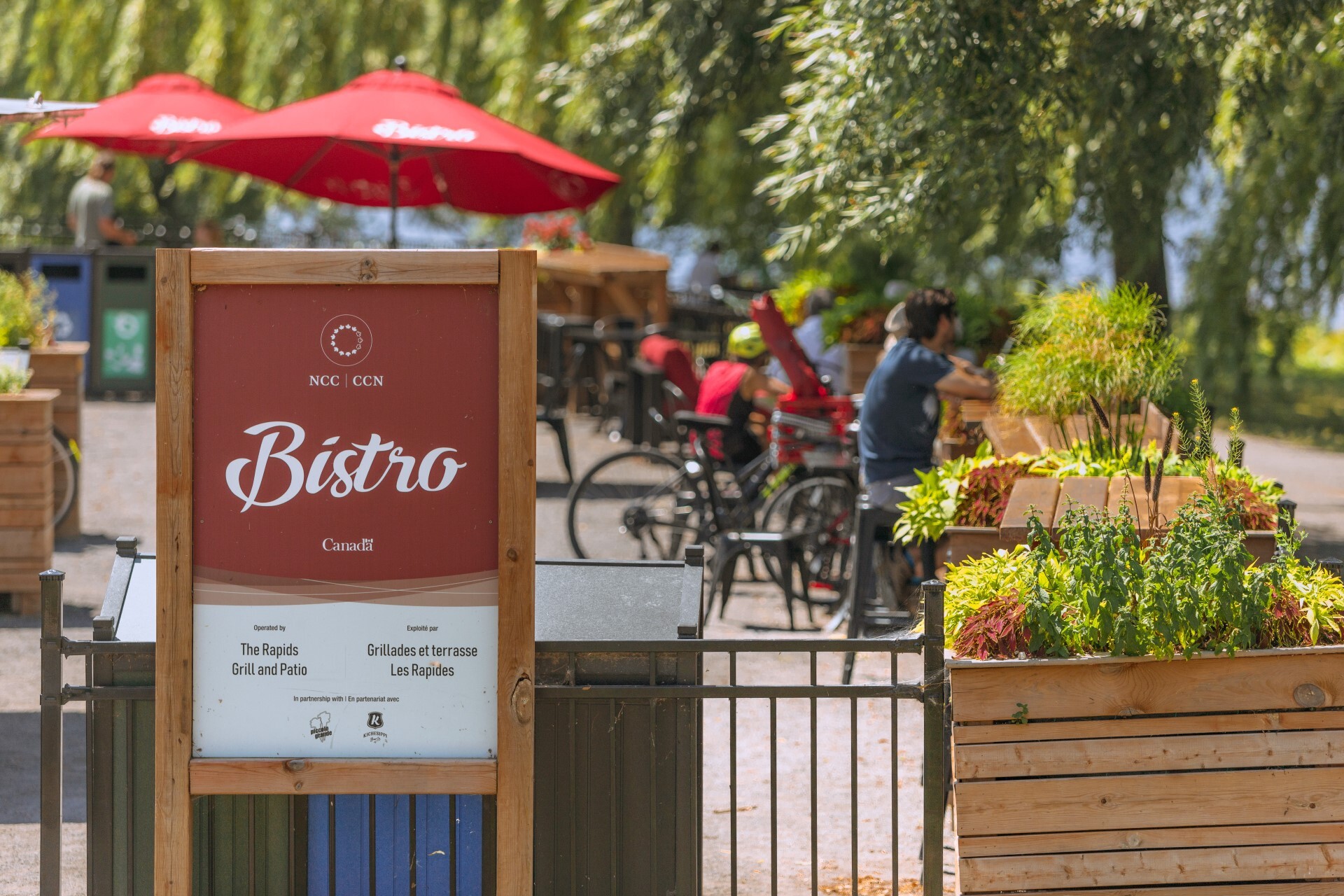 Une enseigne indiquant « Bistro » à gauche et des pots garnis de verdure à droite. Des arbres et des parasols sur une terrasse en arrière-plan.