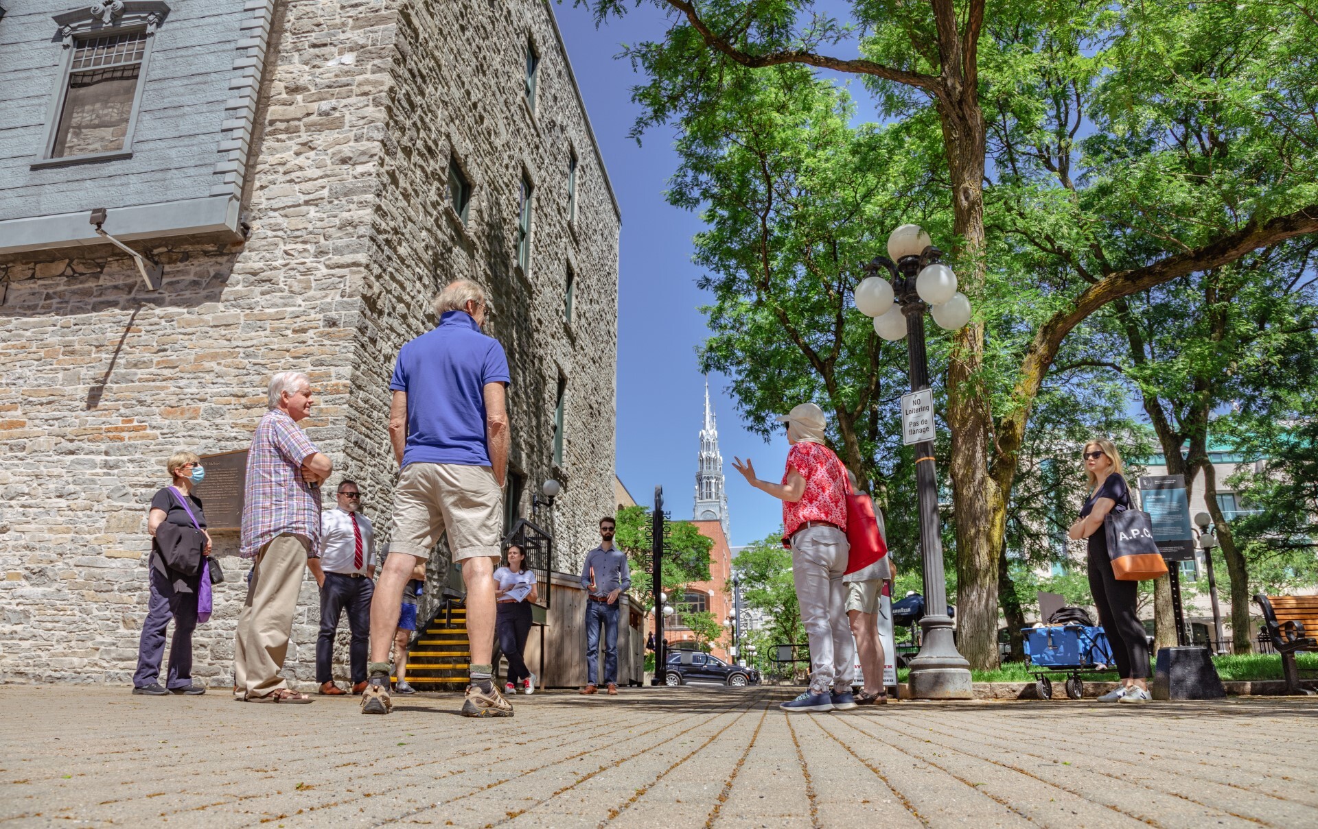 Groupe de personnes écoutant leur guide, en chemise rouge, devant un bâtiment en briques claires.